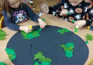Dzieci wyklejają pracę plastyczną. Naklejają skrawki zielonego papieru na kontury kontynentów narysowanych na dużym niebieskim kole symbolizującym kulę ziemską.