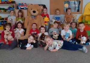 Grupowe zdjęcie Smerfów z misiami. Po między dziećmi duża zabawka misia.Miś jest w kolorze jasnobrązowym i jest dużo więszy od przedszkolaków. Dzieci są bardzo zadowolone, uśmiechnięte i chętnie pozują do zdjęcia.