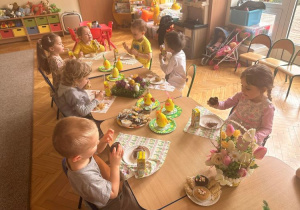 Dzieci siedzą przy stoliku i spożywają poczęstunek wielkanocny, muffinki i soczki przygotowane przez rodziców.