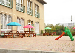 Taras przedszkolny , z kolorową zjeżdżalnią, stolikami drewnianymi i kolorowymi parasolami