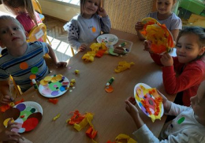Dzieci z grupy Tygrysów siedza przy stoliku w sali przedszkolnej i wykonują prace plastyczną z kółek do origami i bibuły na papierowych talerzykach z okazji Dnia Kropki.