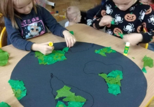 Dzieci wyklejają pracę plastyczną. Naklejają skrawki zielonego papieru na kontury kontynentów narysowanych na dużym niebieskim kole symbolizującym kulę ziemską.