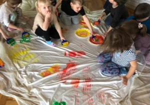 Dzieci z grupy Żabek stempują kreatywnymi wałeczkami płachtę białego materiału, używając kolorowych farb.