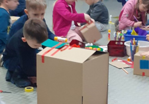 Dzieci tworzą kartonowe budowle używając kolorowych taśm i różnorodnych materiałów plastycznych.