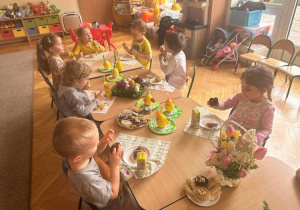 Dzieci siedzą przy stoliku i spożywają poczęstunek wielkanocny, muffinki i soczki przygotowane przez rodziców.