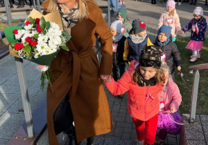 Dzieci z nauczycielką składają kwiaty pod tablicą upamiętniajacą Rocznicę Odzyskania Niepodległości przez Polskę.
